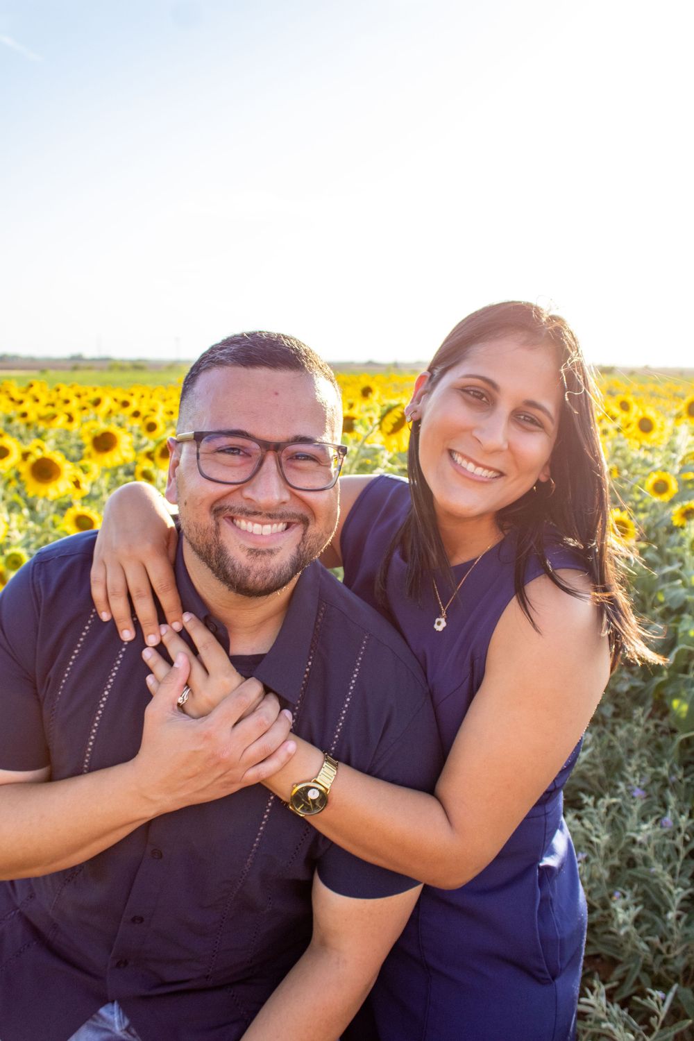 A smiling couple in matching navy outfits poses together in a vibrant sunflower field on a sunny day.