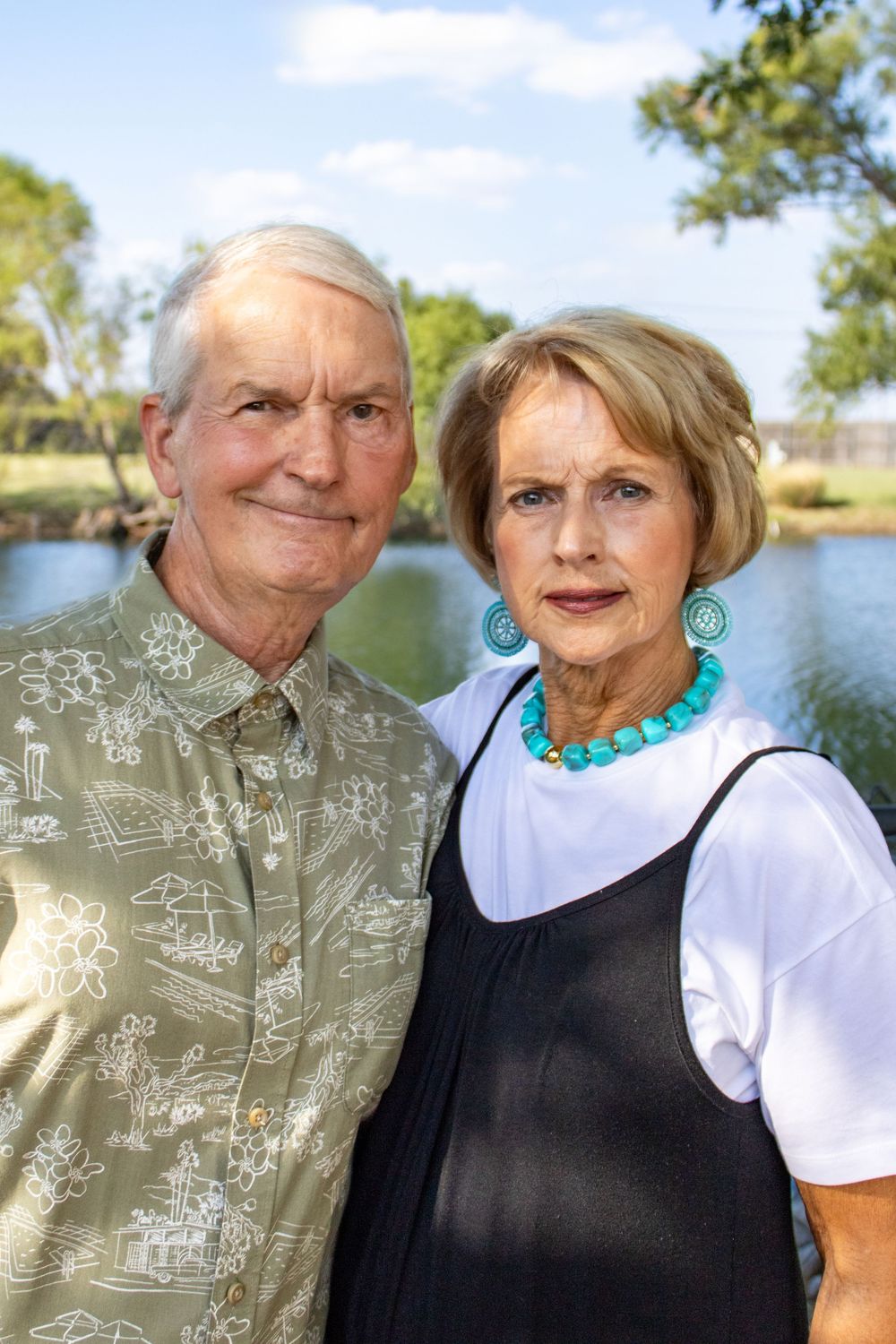 Elderly couple posing outdoors near a pond, woman wearing turquoise jewelry, man in tropical print shirt.