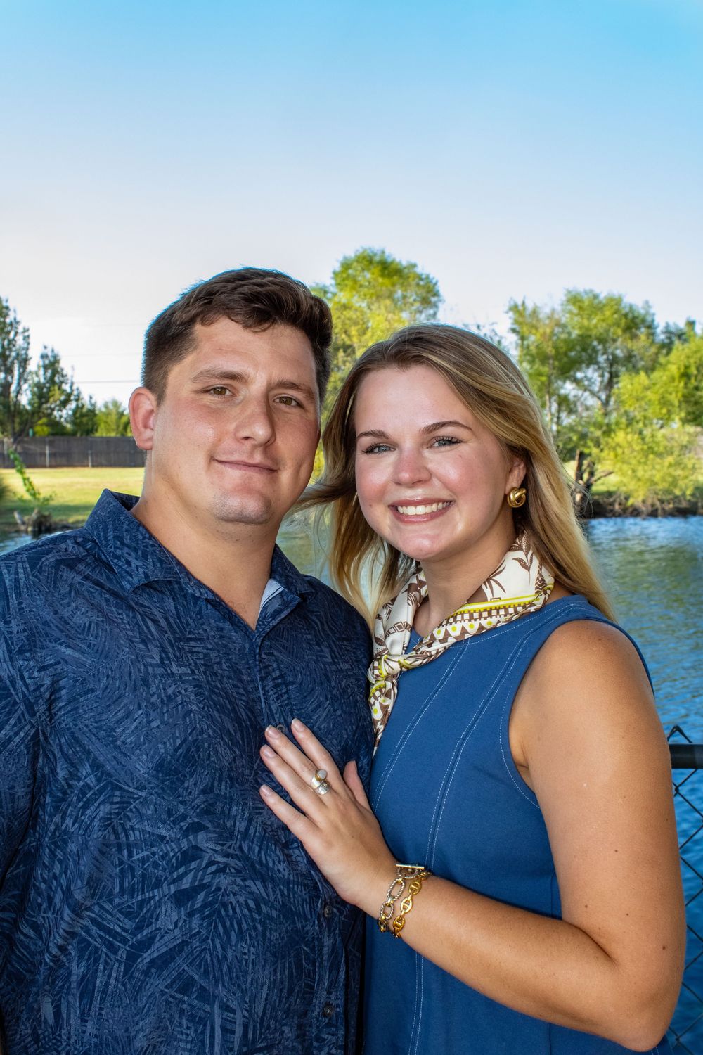Smiling couple in blue outfits posing outdoors near a pond with green trees in the background on a sunny day.