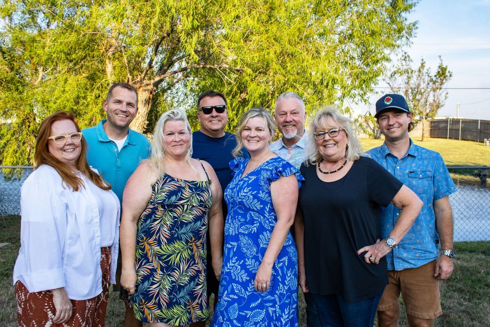 A group of eight smiling adults posing together outdoors at a casual gathering during golden hour.