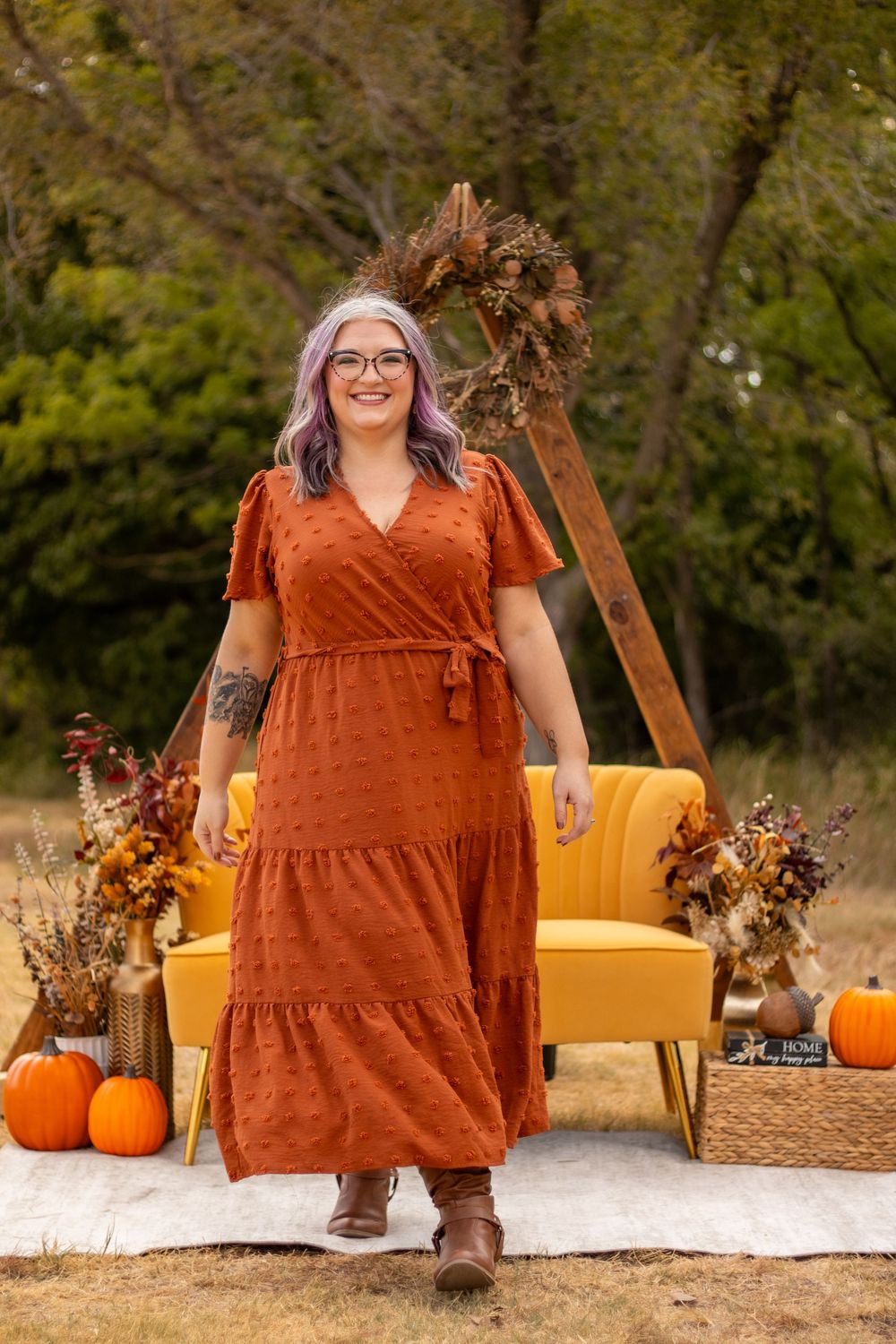 Woman in rust wrap maxi dress walks outdoors among pumpkins and fall decor with yellow chair backdrop.