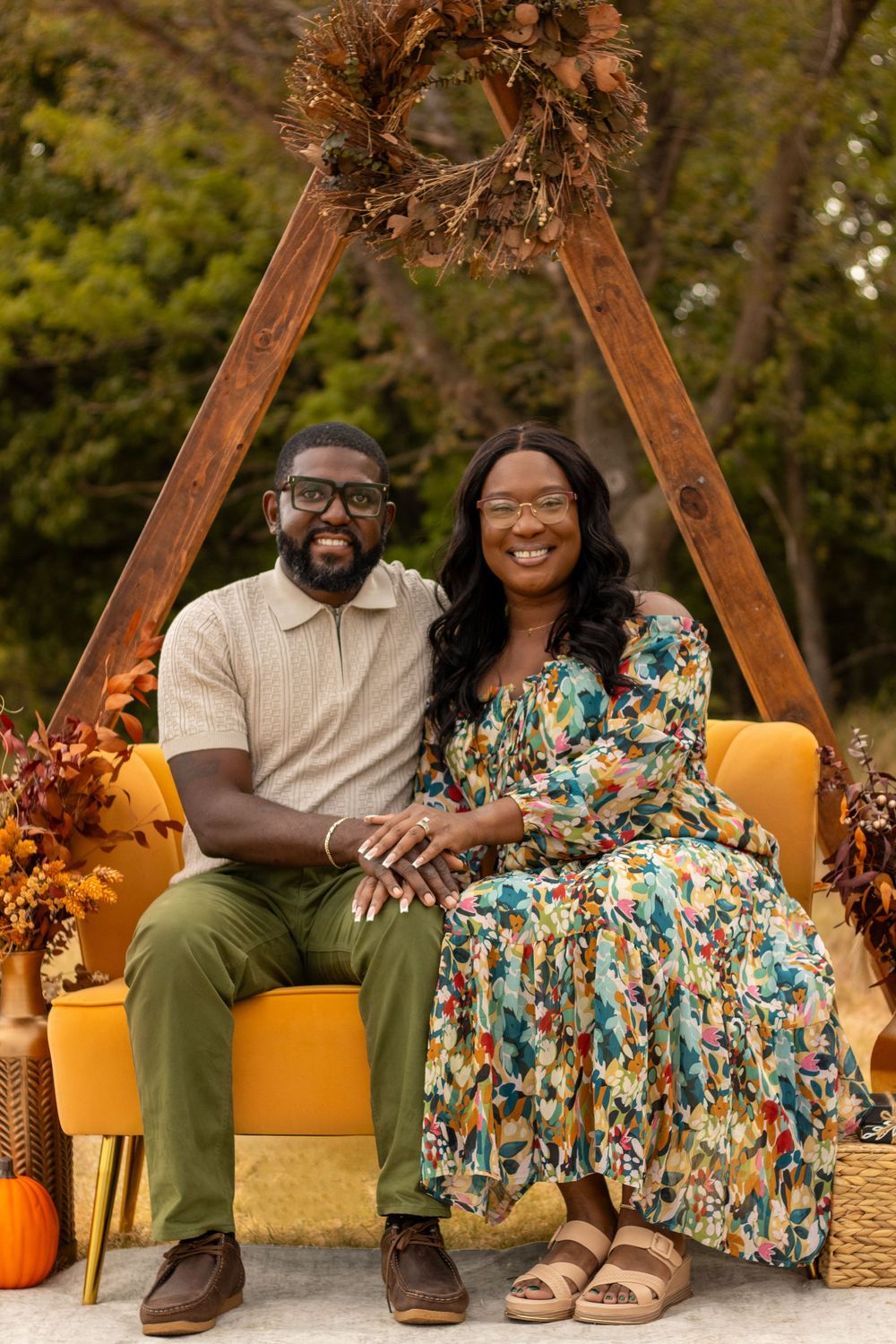 A smiling couple sits together on a yellow sofa outdoors, surrounded by fall decor, pumpkins, and autumn floral arrangements.