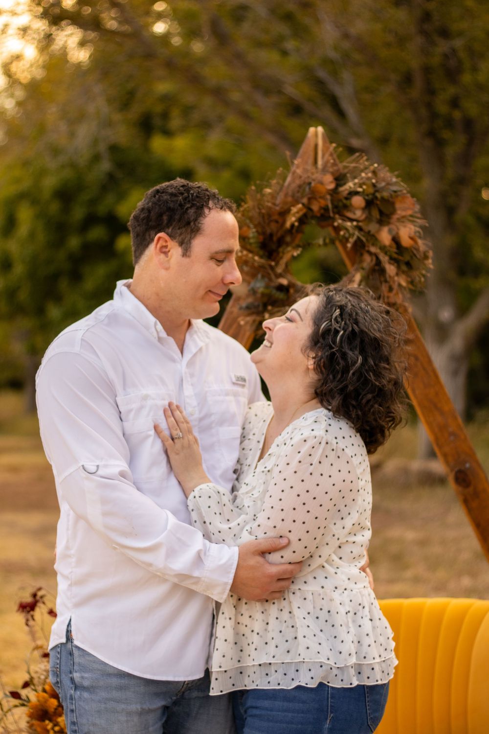 Couple embracing and laughing outdoors during golden hour with a rustic floral arch and yellow velvet chair.