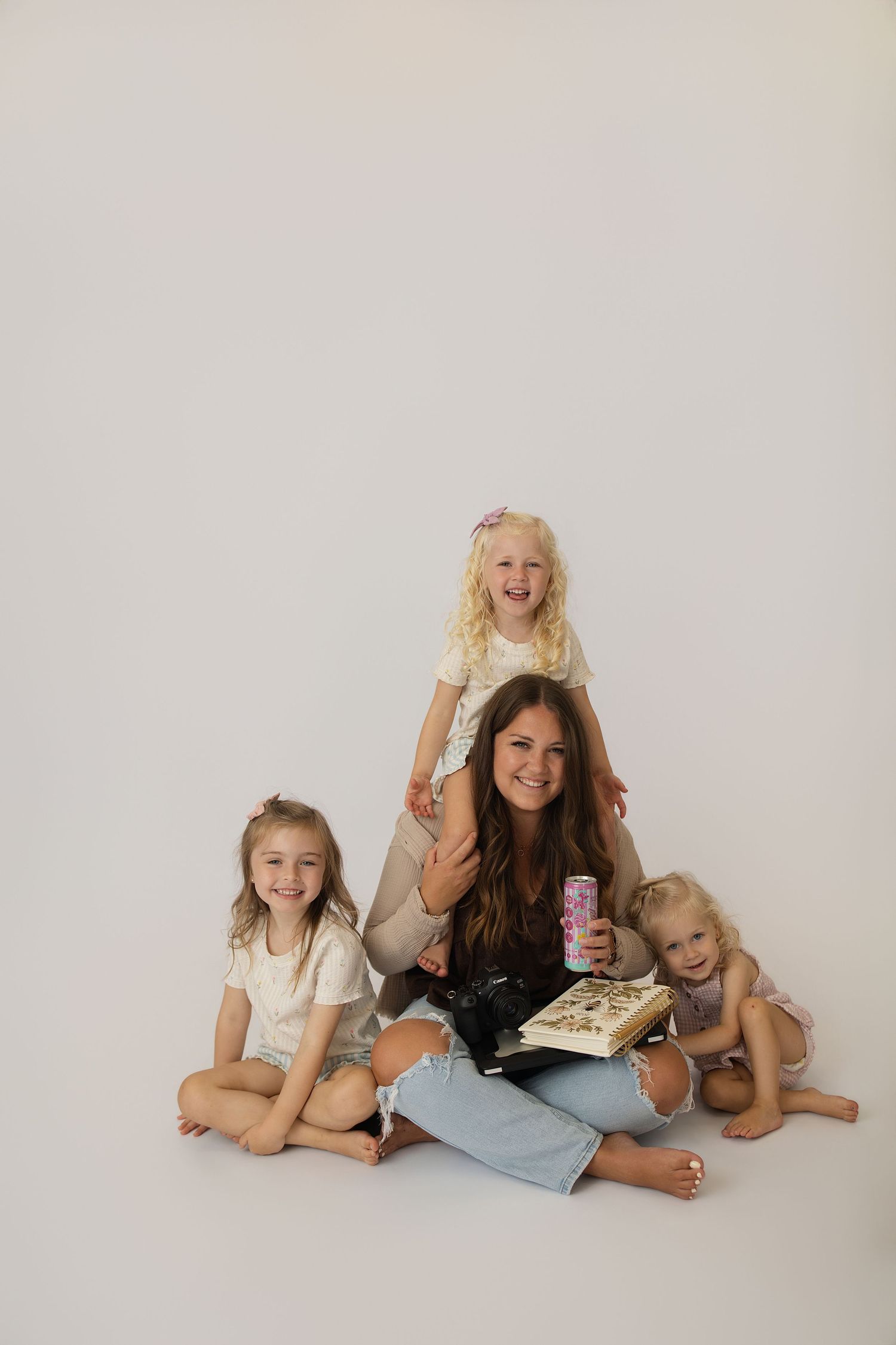A smiling woman sits cross-legged on a white background surrounded by three young blonde girls, holding a book and a drink.