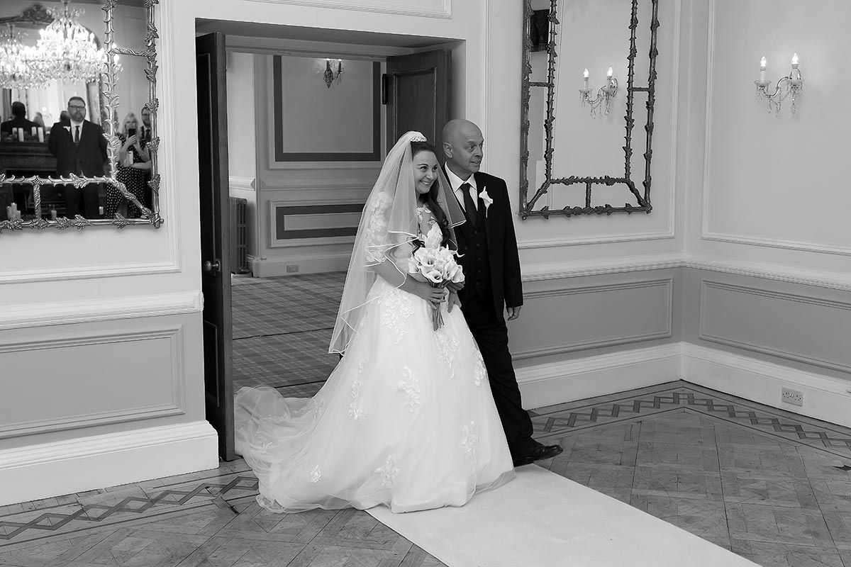 Black and white photo of a bride in a white gown and veil walking with her father at Auchen Castle