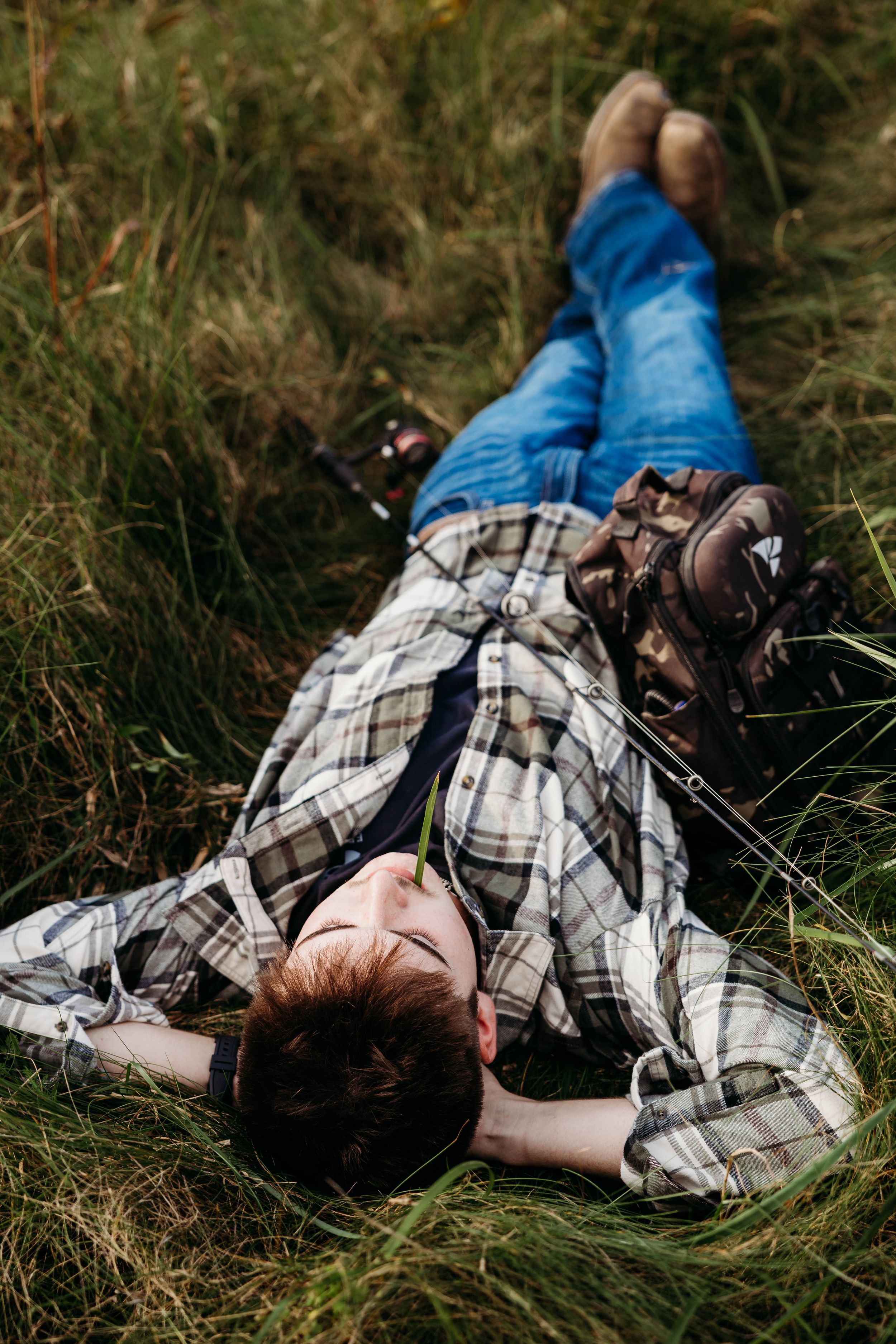 Young man in a plaid flannel shirt and jeans lying back in tall green grass with a blade of grass in his mouth, shot from above.