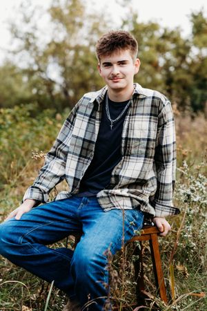 Young man in a plaid flannel shirt and jeans sitting on a wooden stool in a grassy field during an outdoor senior portrait session.
