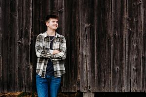 Smiling young man in a plaid flannel shirt and jeans stands with arms crossed against a weathered dark wooden barn wall.