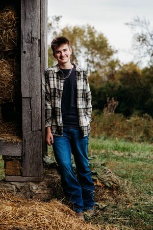 Smiling young man in a plaid flannel jacket and jeans leaning against a rustic wooden barn with hay bales and autumn trees behind him.