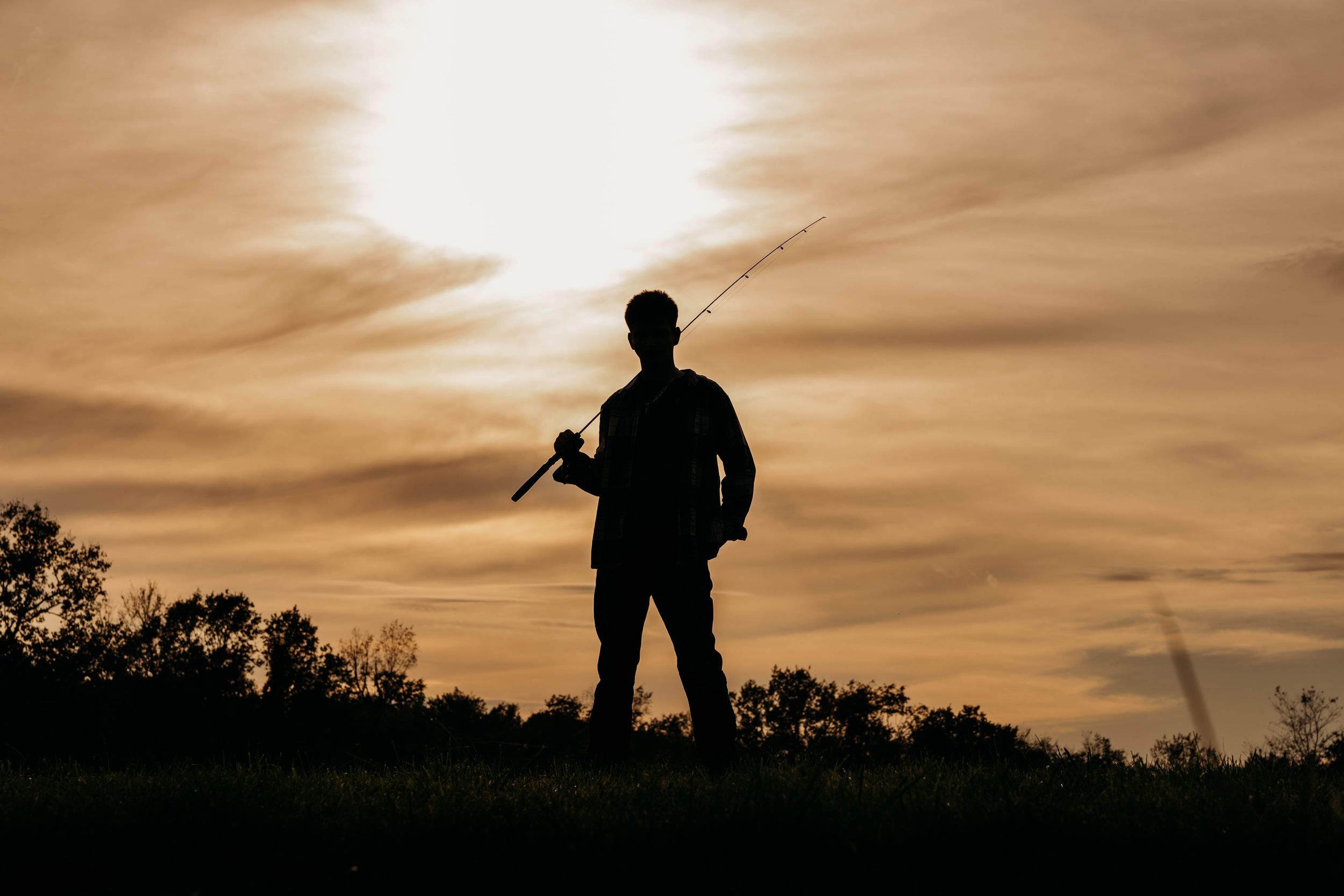 Silhouette of a man holding a fishing rod over his shoulder against a golden sunset sky with soft clouds above a dark treeline.