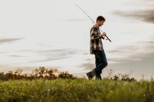 Man in a plaid flannel shirt and jeans walks through a grassy field at dusk carrying a fishing rod under a soft, cloudy sky.