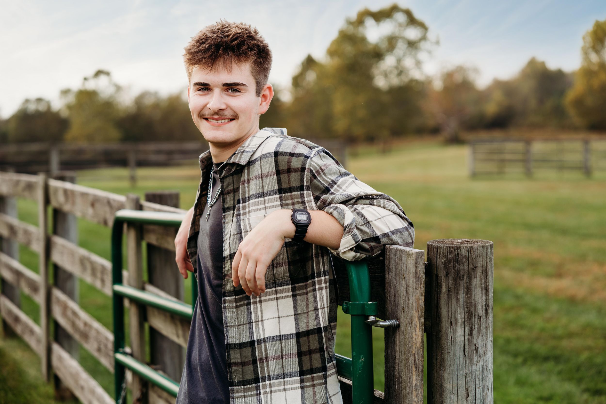Smiling young man in a plaid flannel shirt leans on a wooden and green metal farm fence with a grassy pasture and trees behind him.