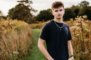 Young man in a black t-shirt with a cross necklace stands on a grassy path surrounded by tall golden wildflowers and trees at sunset.