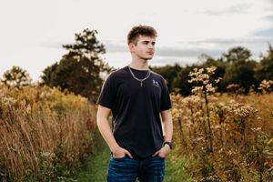 Young man in a black t-shirt and jeans stands with hands in pockets on a grassy path surrounded by tall golden wildflowers at sunset.