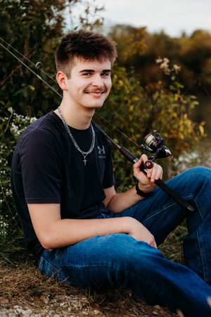Smiling young man in a black t-shirt and jeans sits on a grassy bank holding a fishing rod, with autumn foliage in the background.
