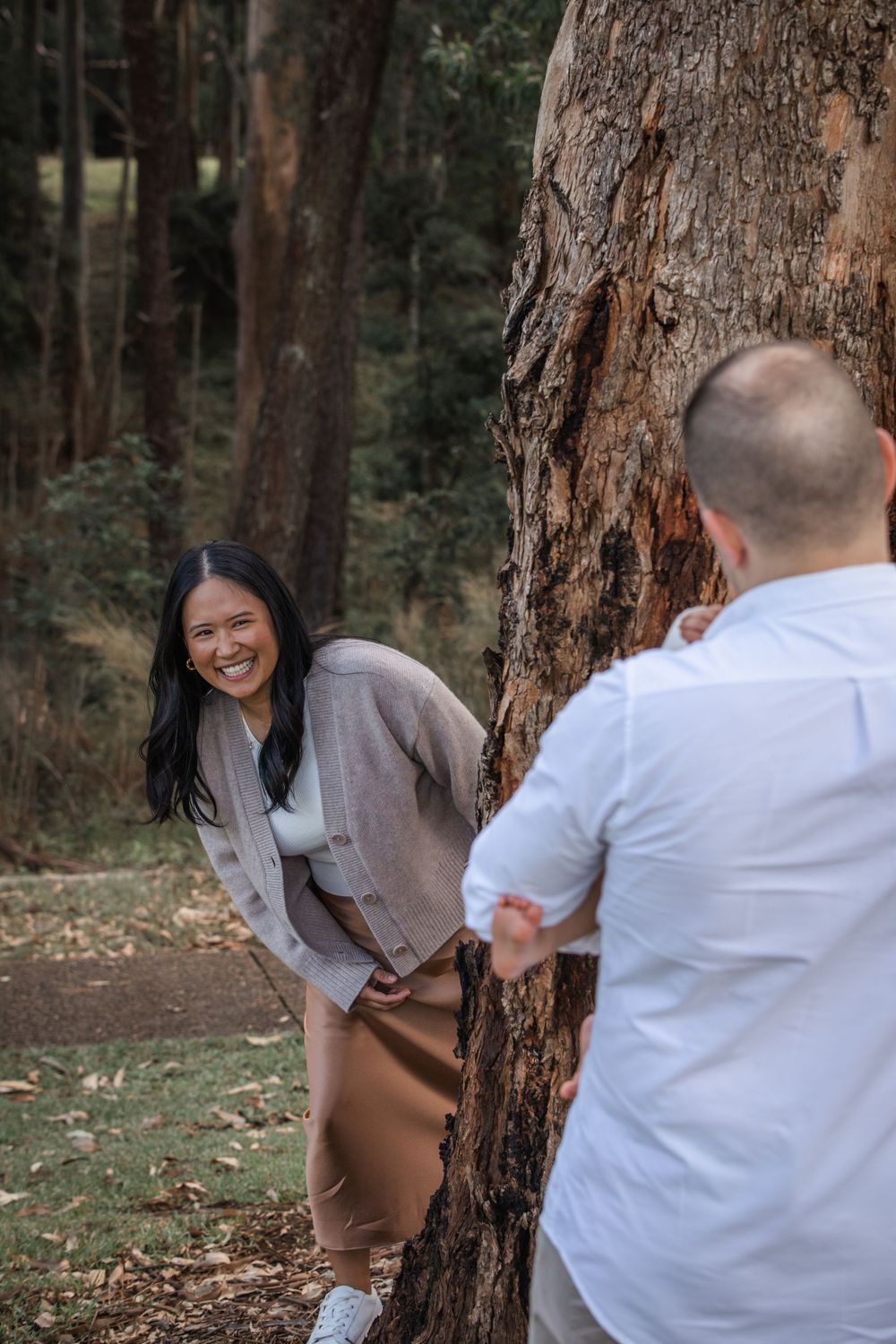 Smiling woman in a beige cardigan peeks playfully around a large tree trunk at a man in a white shirt in a sunny forest in Castle Hill.