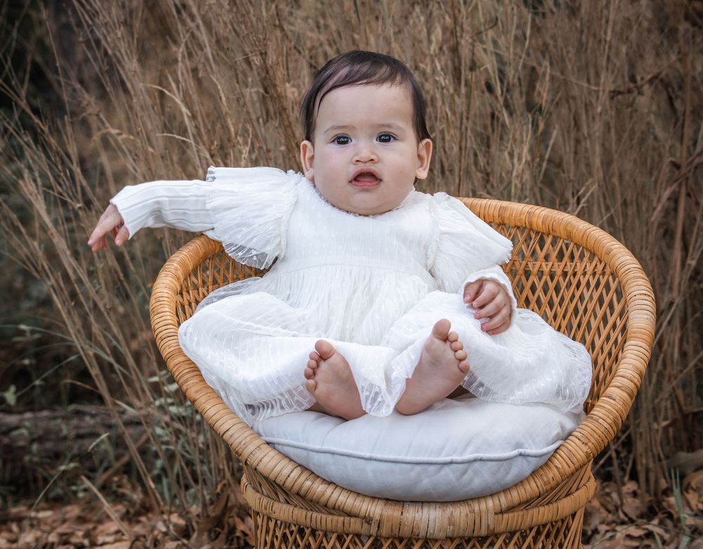 Baby in a white long-sleeve dress sits in a round wicker chair with a cushion, surrounded by tall dry grass at Castle Hill Heritage Park.