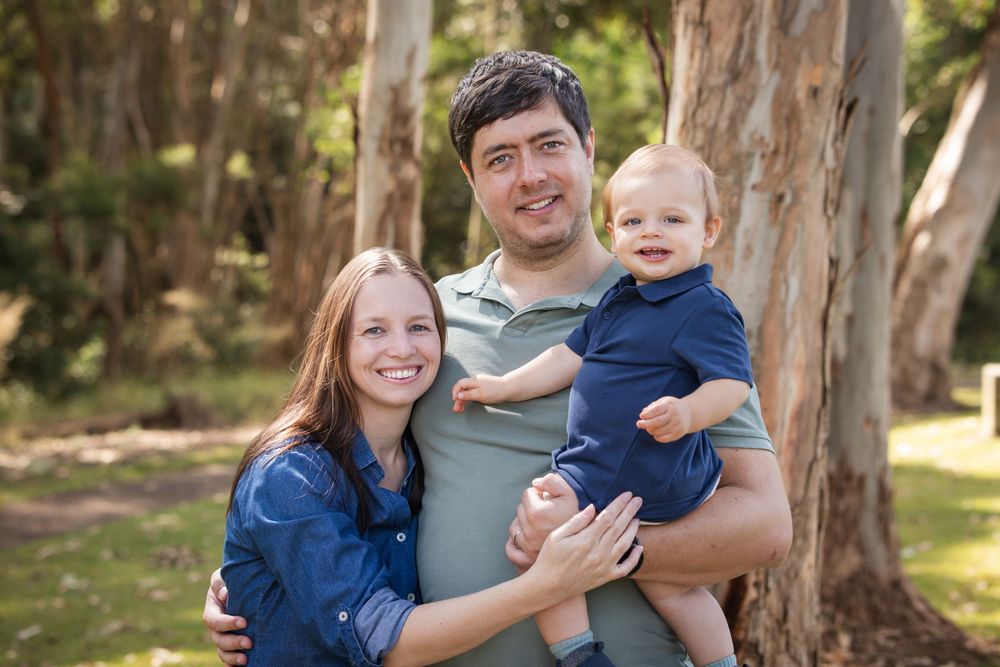 Smiling family of three posing outdoors among tall eucalyptus trees, with a woman and man holding a toddler in a blue outfit in Castle Hill.