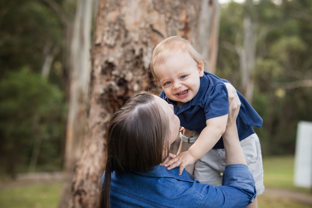 Smiling toddler in a navy shirt is lifted by a woman in a denim top beside a large tree trunk in a sunny Castle Hill Heritage Park.
