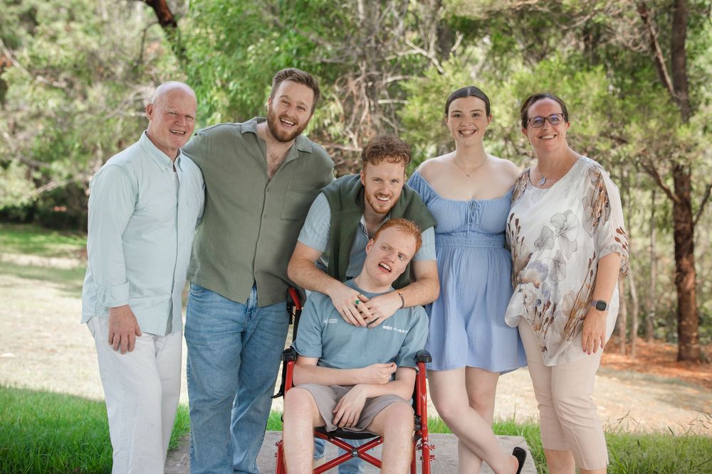 Smiling family of six poses outdoors among green trees, with a young man seated in a red wheelchair and loved ones gathered around him.