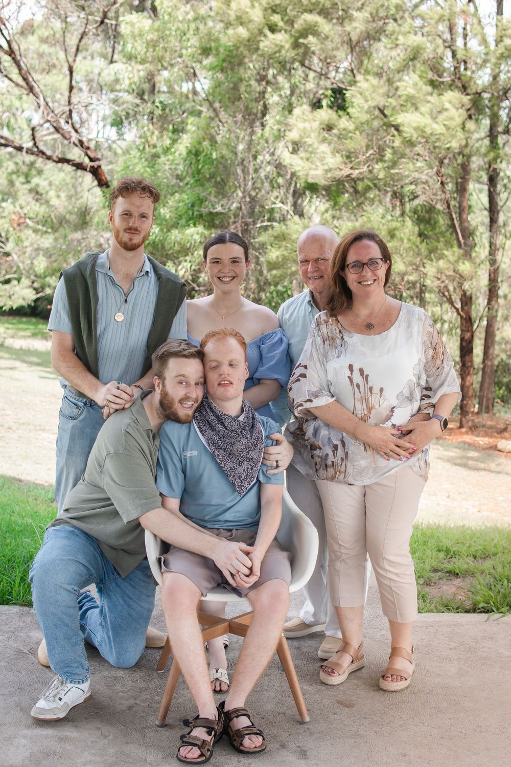 Six family members of different ages pose closely together outdoors on a patio, with green trees and a sunny park setting behind them.