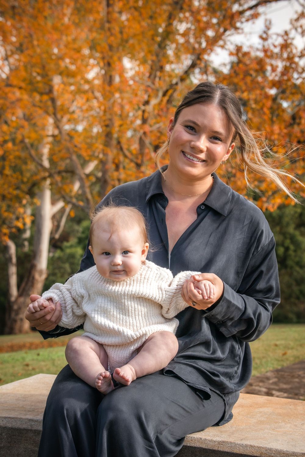 A smiling woman in a dark shirt holds a baby in a cream knit sweater on a stone bench in front of orange autumn trees in Castle Hill.