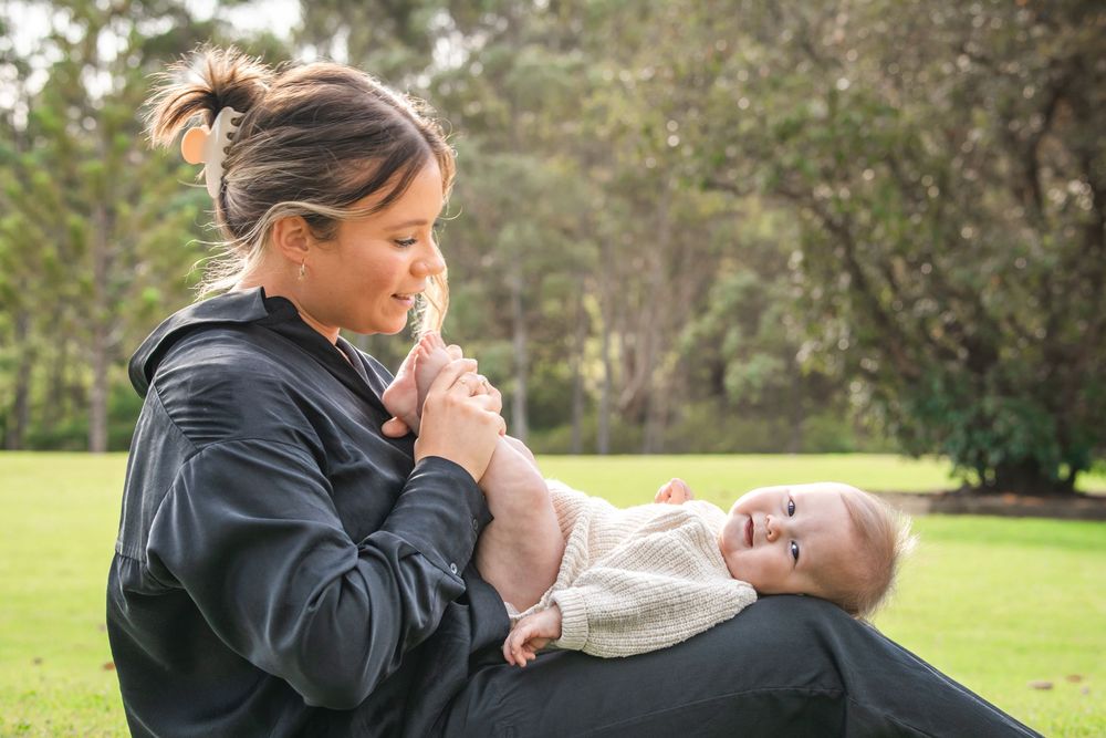 A smiling mother sits on a grassy lawn holding her baby's feet as the infant lies in her lap in a sunny Castle Hill Heritage Park.