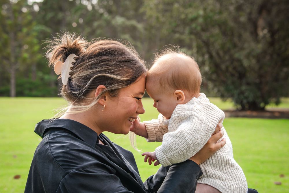A smiling woman in a black jacket touches foreheads with a baby in a knit sweater while standing on a sunny green lawn in Castle Hill.