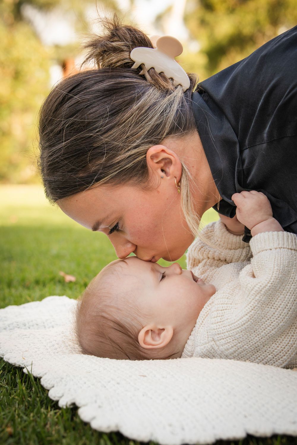 A mother leans down to kiss her baby, who lies on a cream knit blanket on green grass in a sunny at Castle Hill Heritage Park.