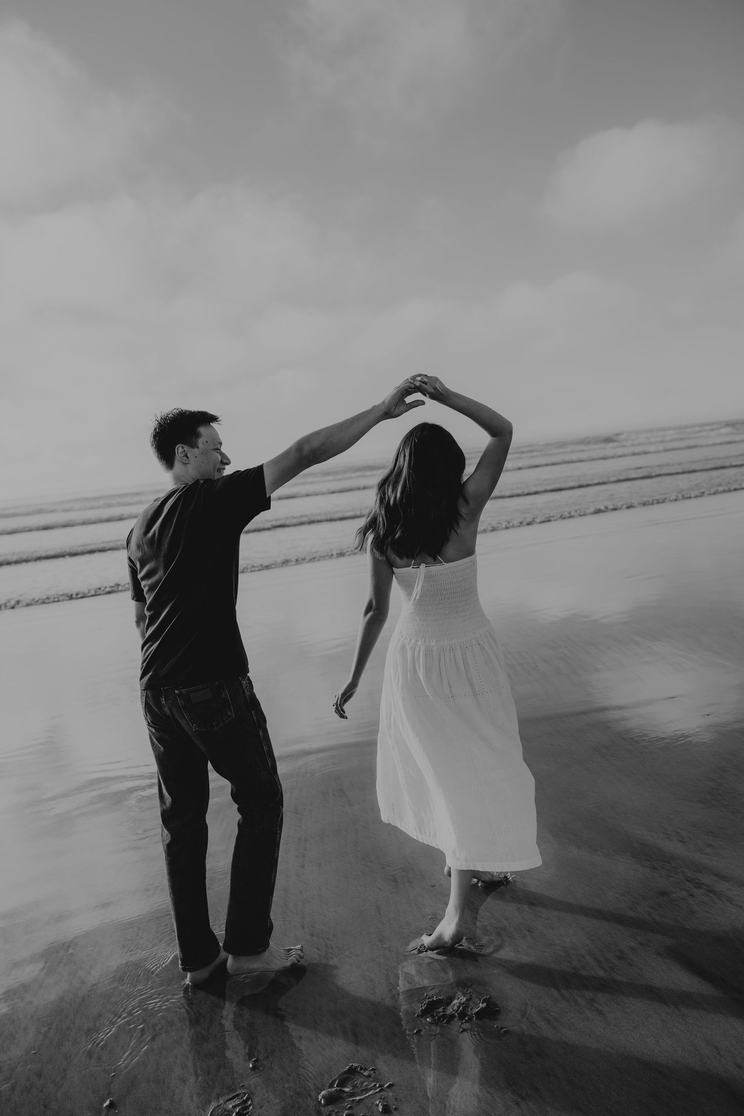 Black and white photo of a couple dancing barefoot on a wet beach, man spinning woman in a white dress.