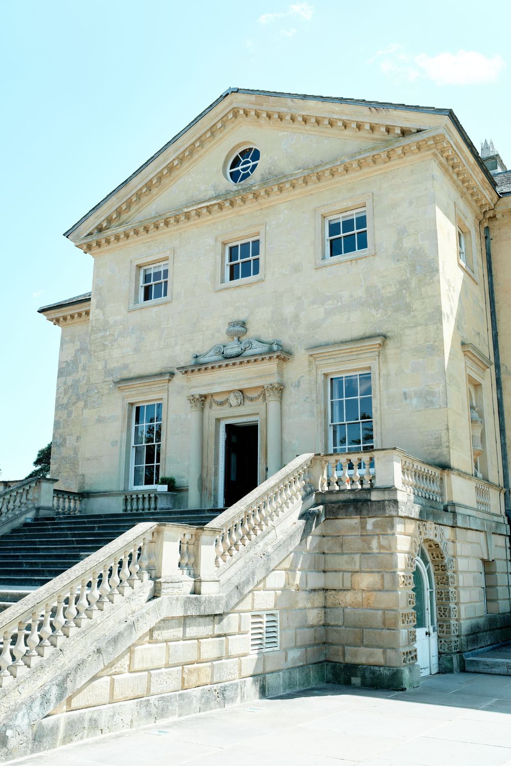 Grand neoclassical stone manor house with ornate balustrade staircase and decorative facade under clear blue sky.