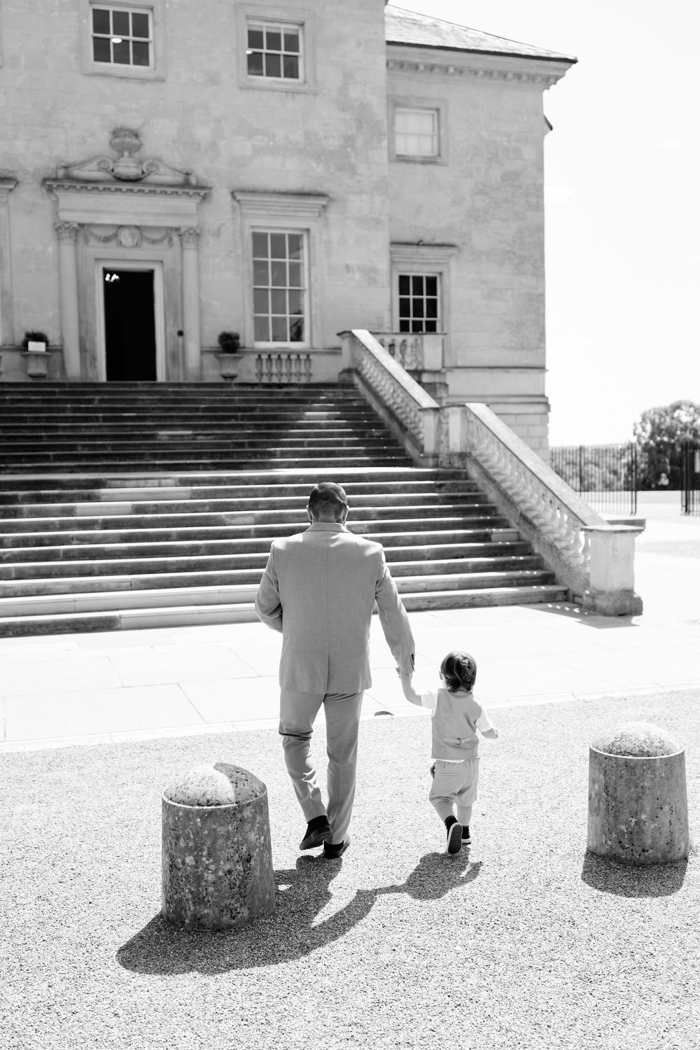 Black and white photo of a man in a suit holding hands with a small child walking toward grand estate steps.