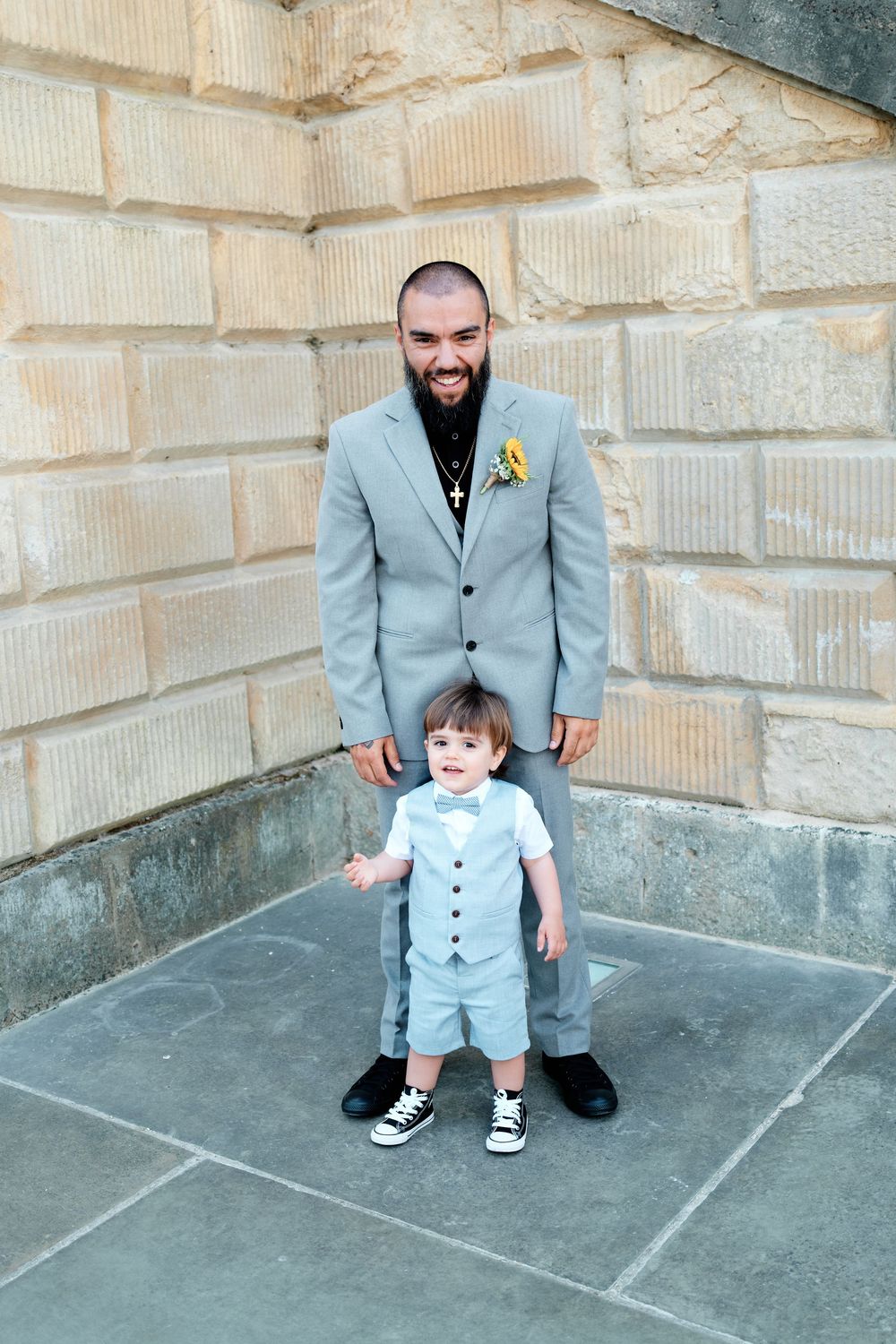 A bearded groom in a gray suit with sunflower boutonniere stands behind a toddler in a light blue outfit with bow tie.
