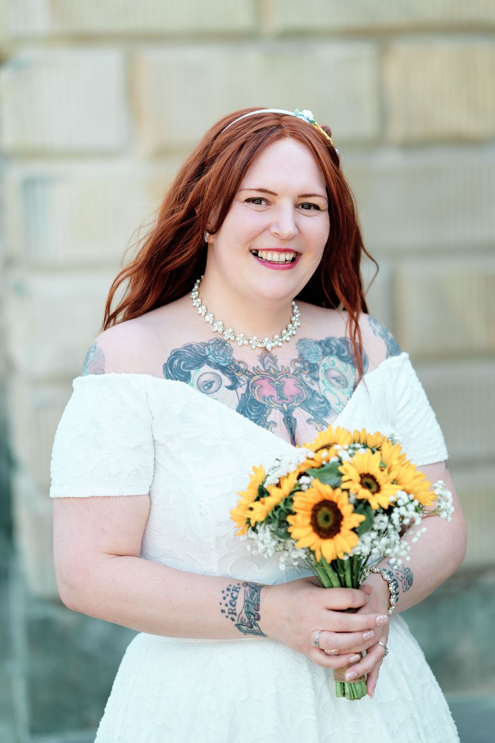 Smiling red-haired bride in white off-shoulder dress holding sunflower bouquet, with chest tattoos and floral headpiece.