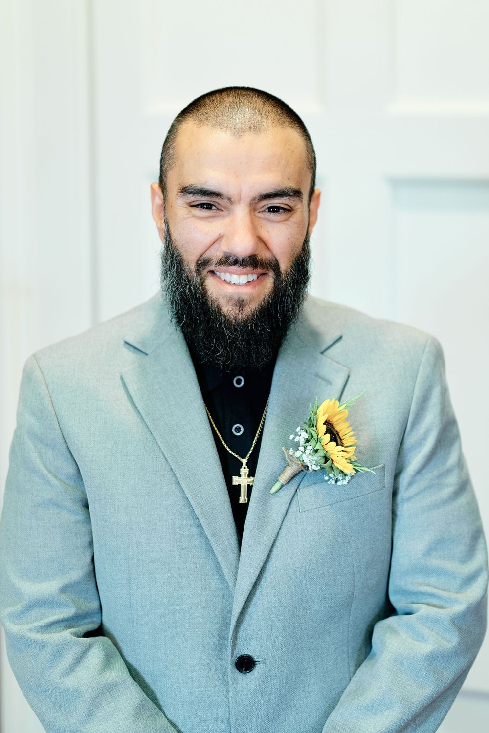 Smiling groom with beard wearing gray suit, cross necklace, and sunflower boutonniere at wedding.