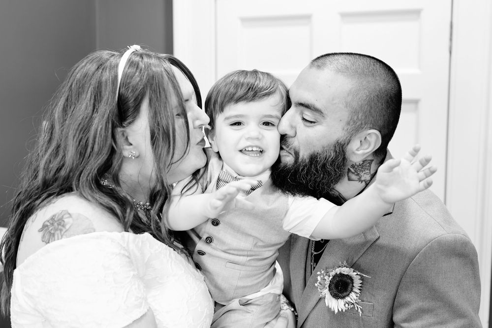 Black and white photo of parents kissing their smiling toddler on the cheeks during a wedding celebration.