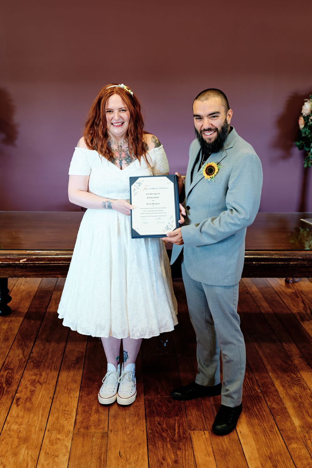 Couple in wedding attire holding marriage certificate, bride in white dress with red hair, groom in light gray suit with sunflower.