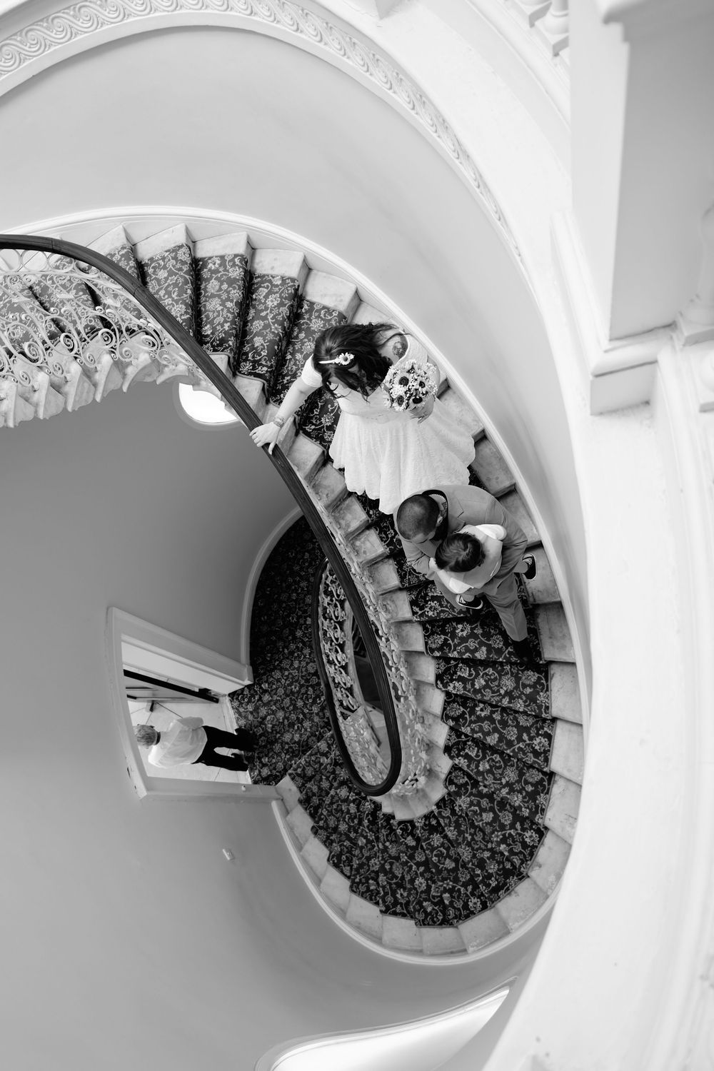 Black and white aerial view of a bride and groom descending an elegant spiral staircase.