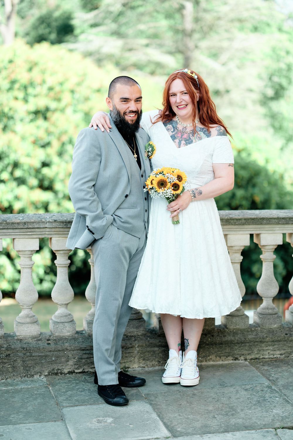 Tattooed bride and groom in gray suit pose with sunflower bouquet on stone balcony at wedding.