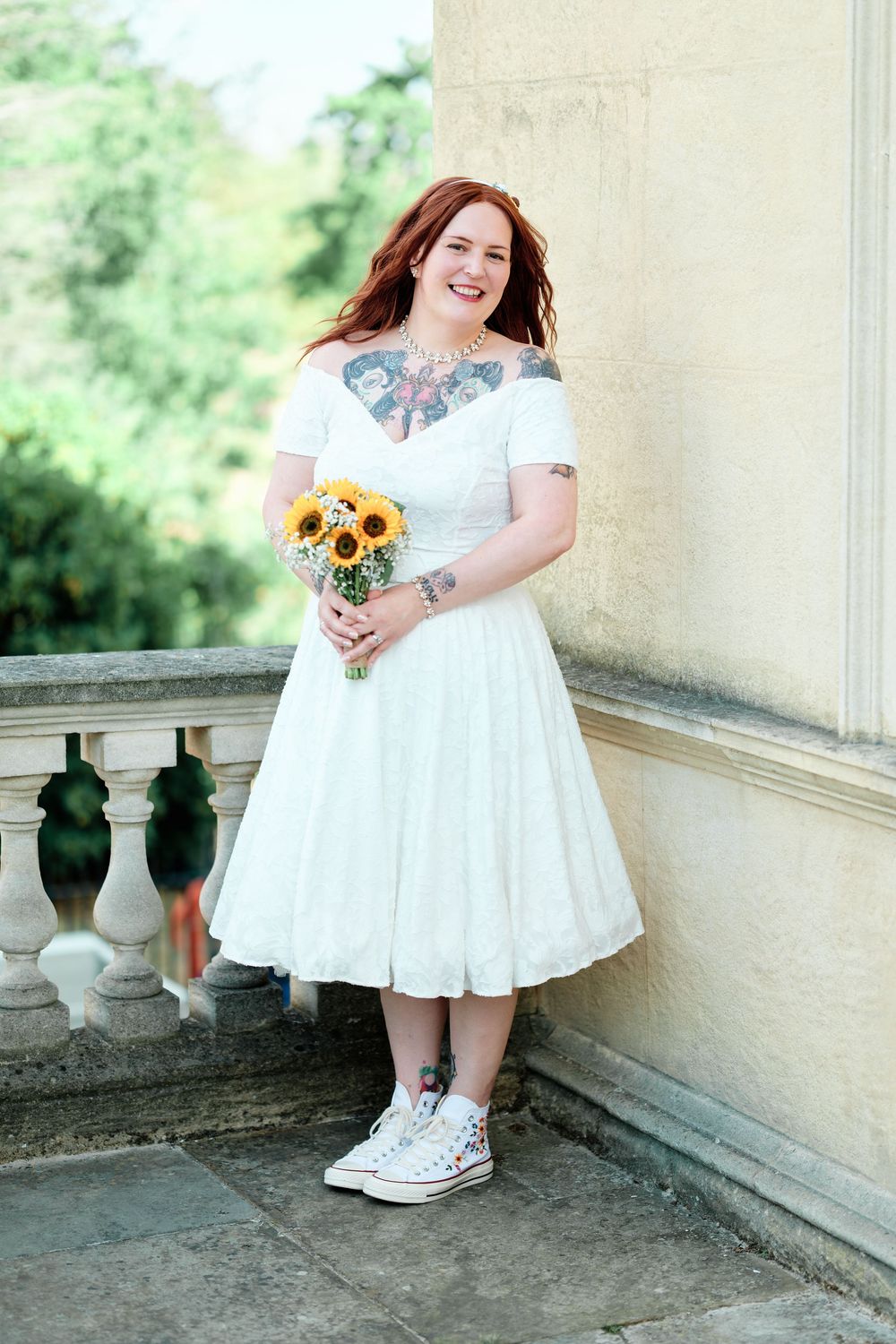 Tattooed bride in white tea-length dress and Converse sneakers holds sunflower bouquet on stone balcony.