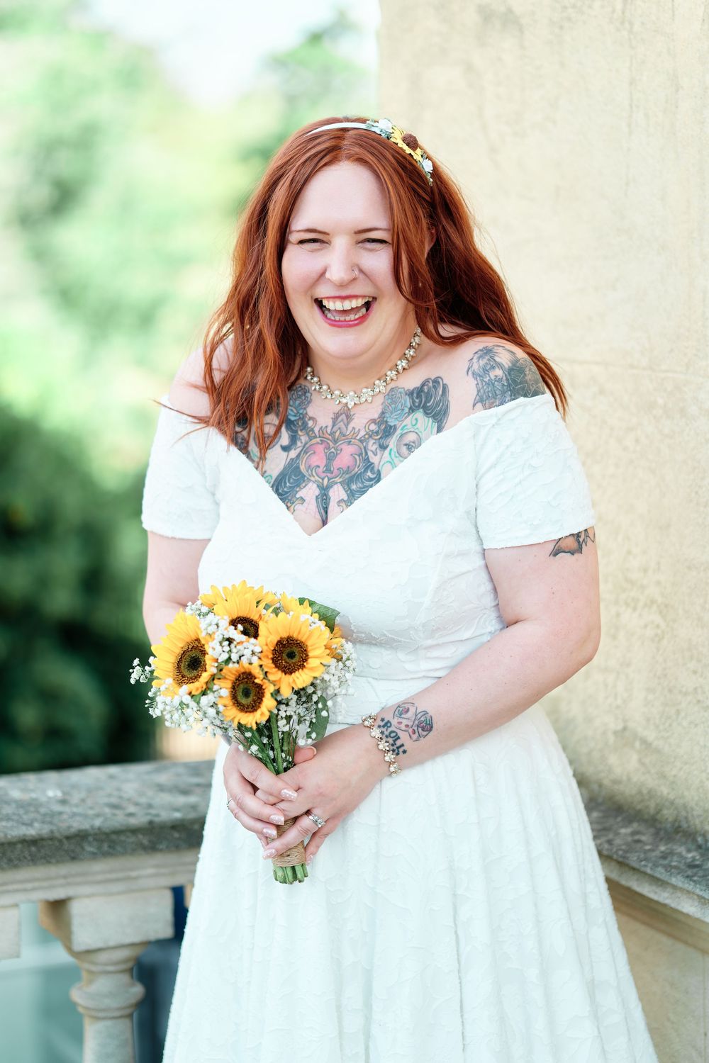 Laughing red-haired bride with tattoos in white dress holding sunflower bouquet at outdoor wedding.
