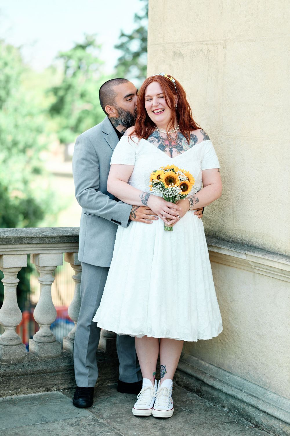 Bride in white dress holding sunflower bouquet laughs as groom in gray suit kisses her cheek on a balcony.