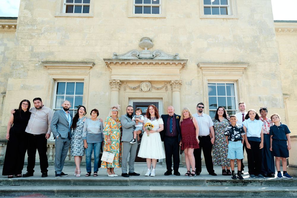 Wedding group photo with bride and groom surrounded by guests on steps of elegant Georgian manor house.