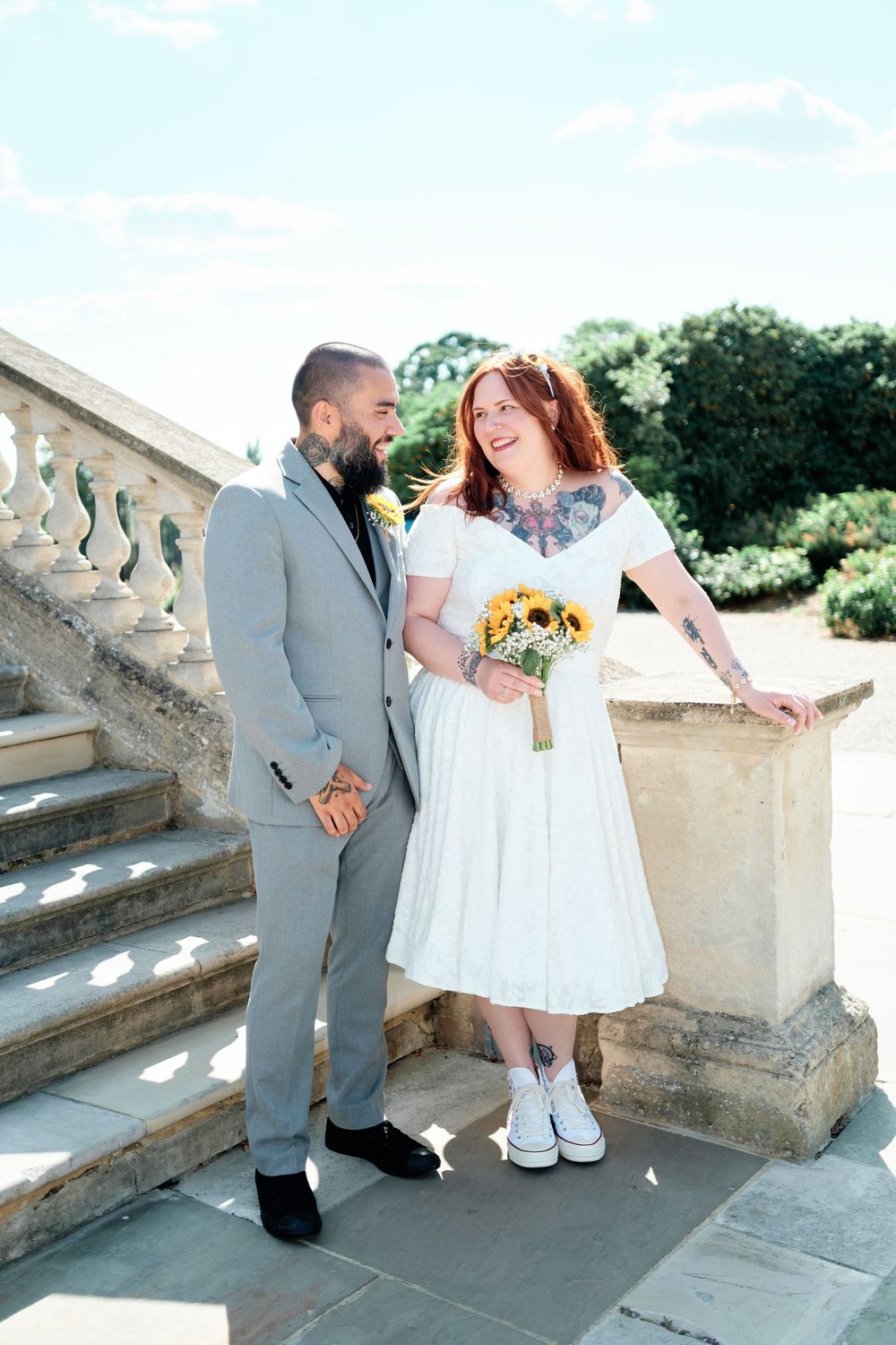 Bride in white dress with sunflowers and groom in gray suit share a smile on stone steps outdoors.