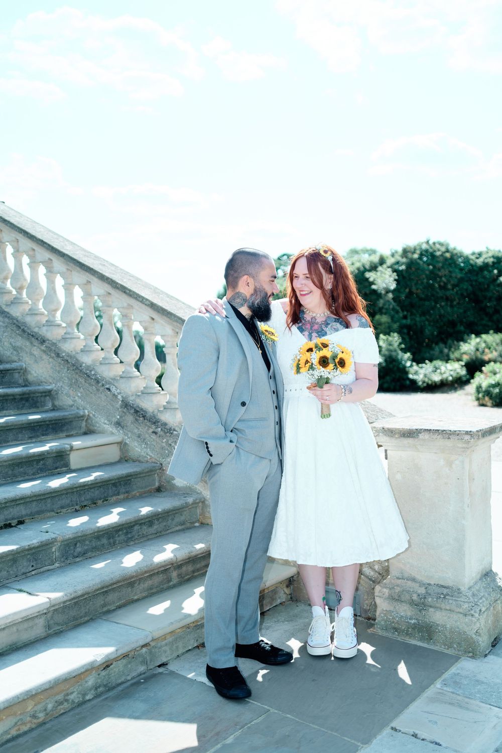 Bride and groom pose on stone steps, she holds sunflower bouquet in white dress, he wears grey suit.