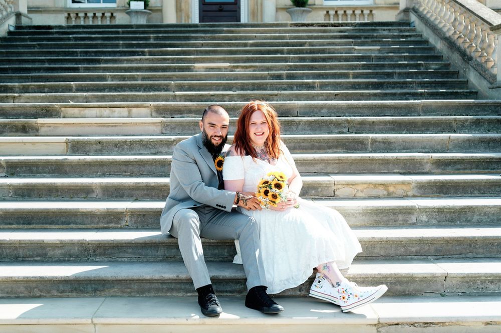Bride and groom sitting on stone steps, bride wearing white dress and Converse shoes, holding sunflower bouquet.
