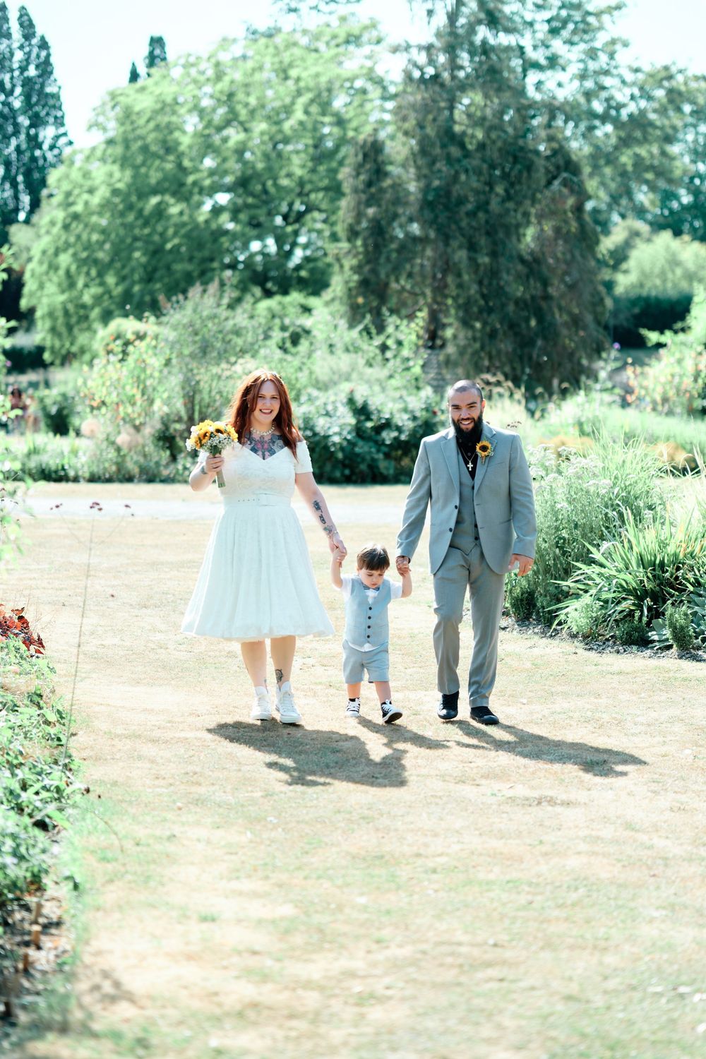 A couple in wedding attire walks with their toddler through a sunny garden path, holding hands and carrying sunflowers.