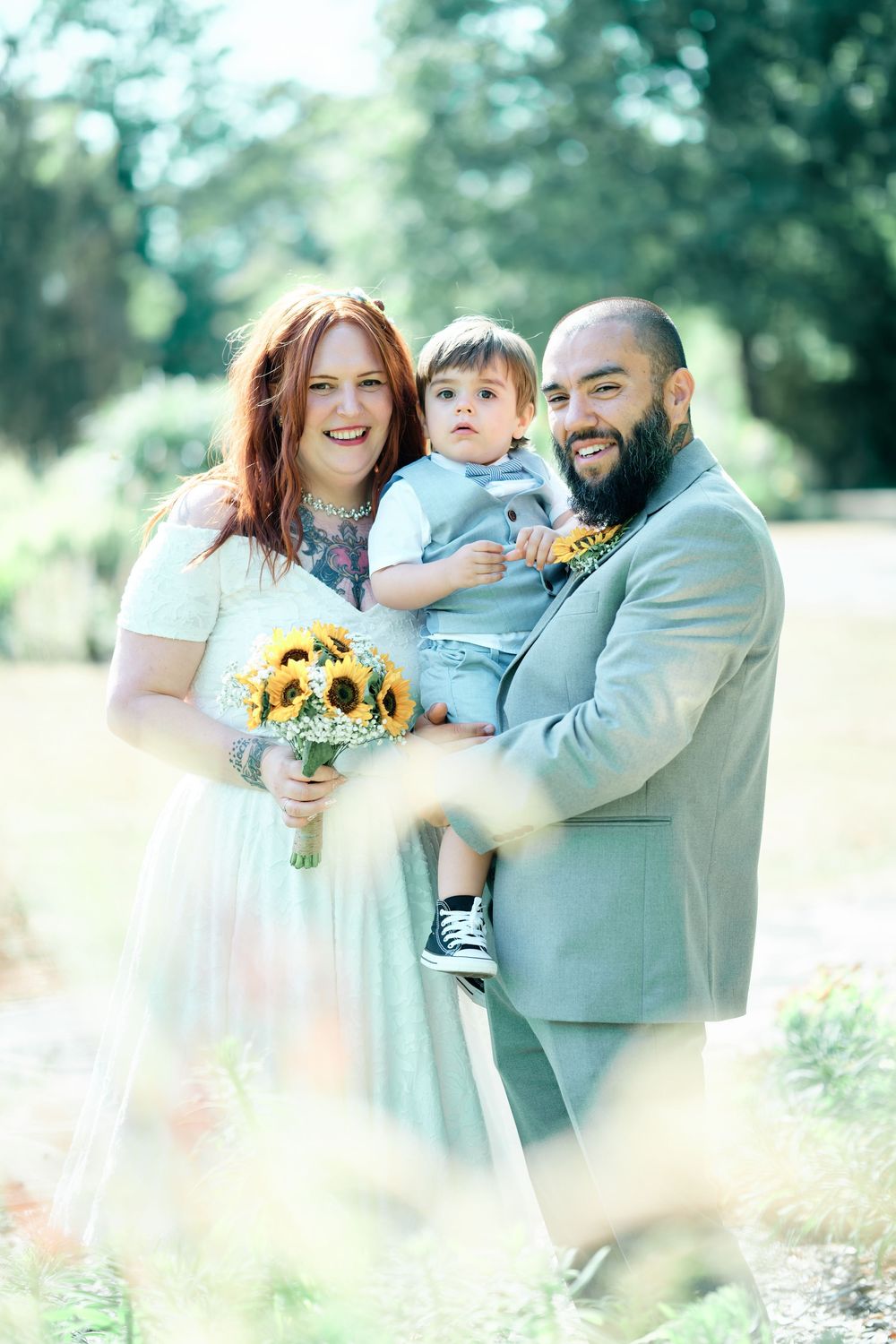 Smiling couple in wedding attire holding toddler outdoors, bride with sunflower bouquet, groom in gray suit with beard.