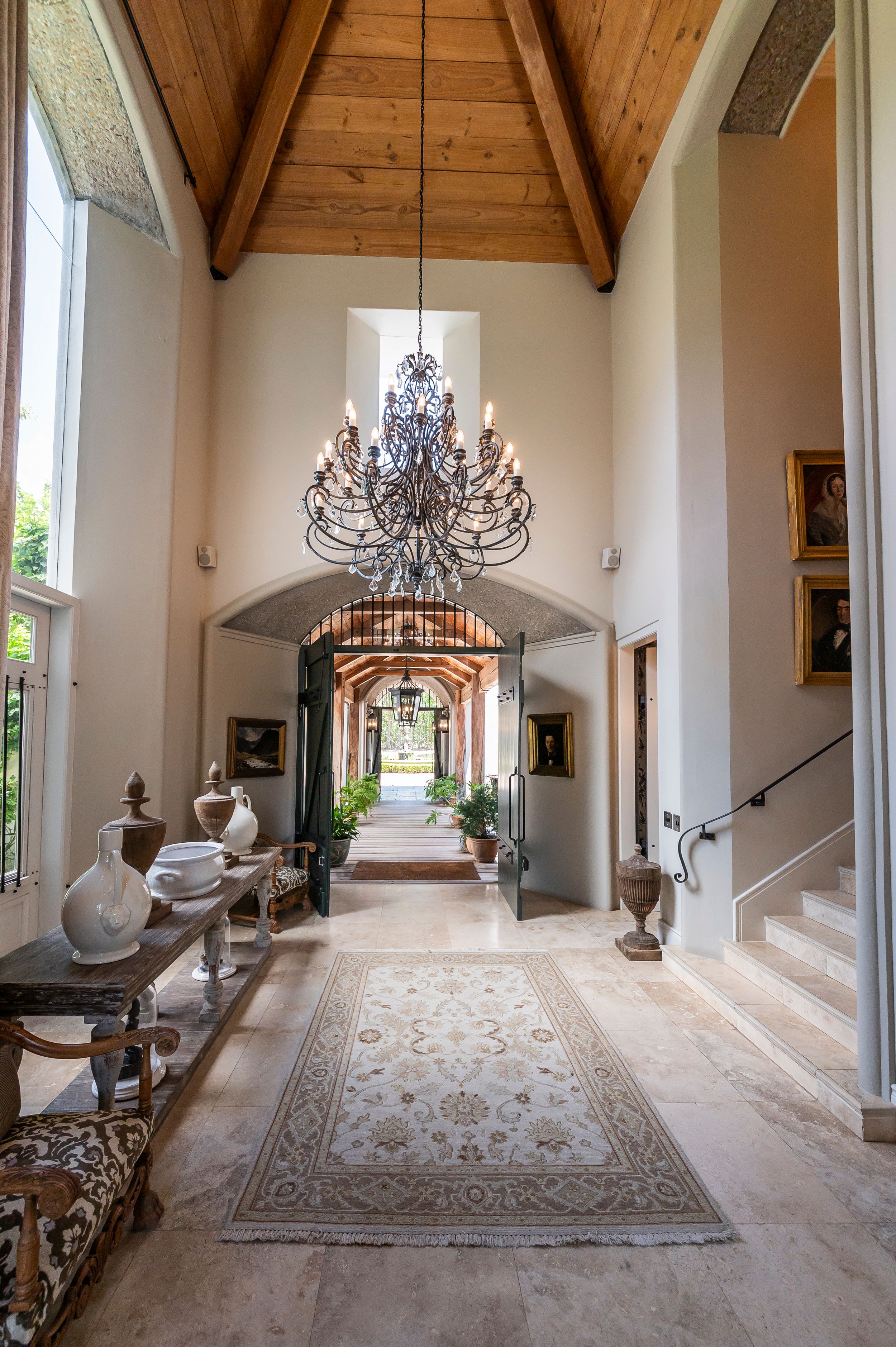 Grand entry hallway with vaulted wood ceiling, wrought iron chandelier, patterned area rug, antique console table and a stone staircase.