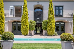 Elegant stone mansion with arched entryways, tall cypress trees, potted shrubs, and a rectangular swimming pool in the foreground.